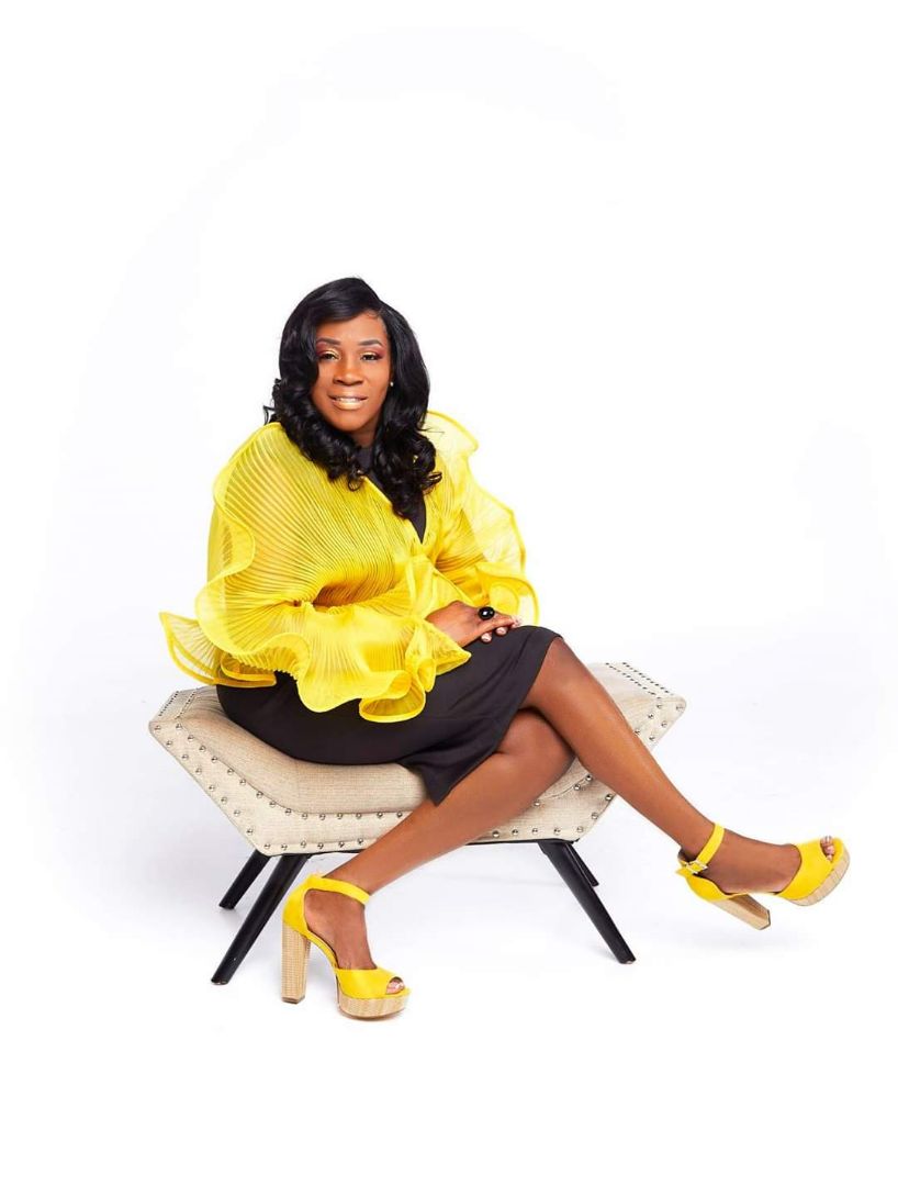 Picture of Carolyn Lewis sitting on a beige bench wearing yellow blouse, black skirt, yellow wedge heels.